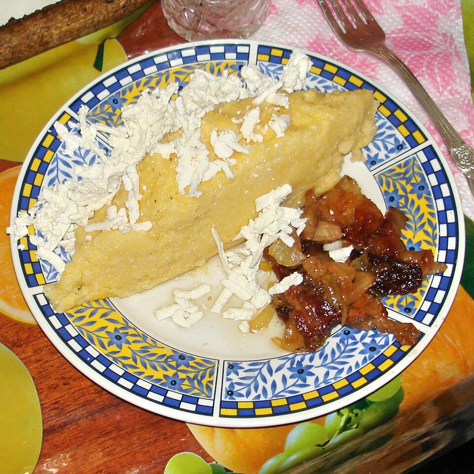 A wedge of mămăligă on a plate, topped with grated cheese, next to fried pork rind